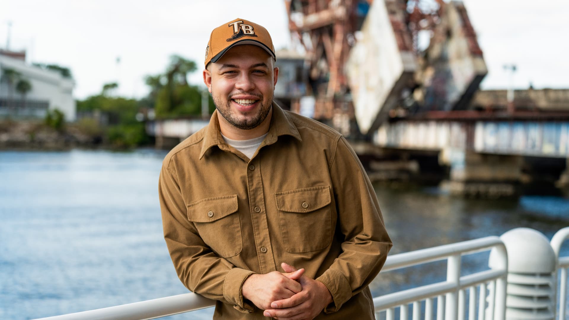 Portrait of a smiling man in a tan work shirt and ball cap leaning on a railing above the Hillsborough River