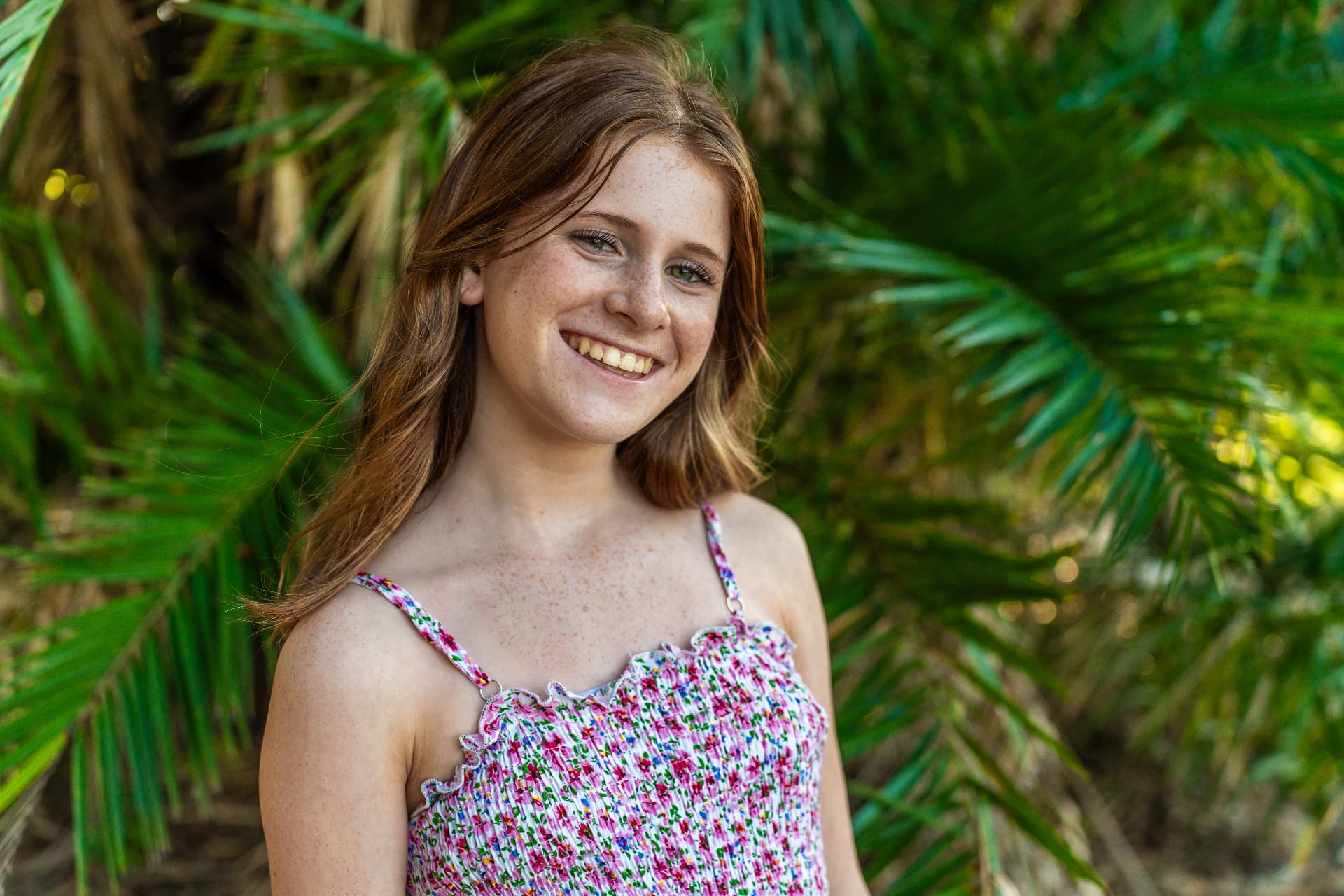 Portrait of a freckled young woman with auburn hair smiling in front of sunlit palm fronds