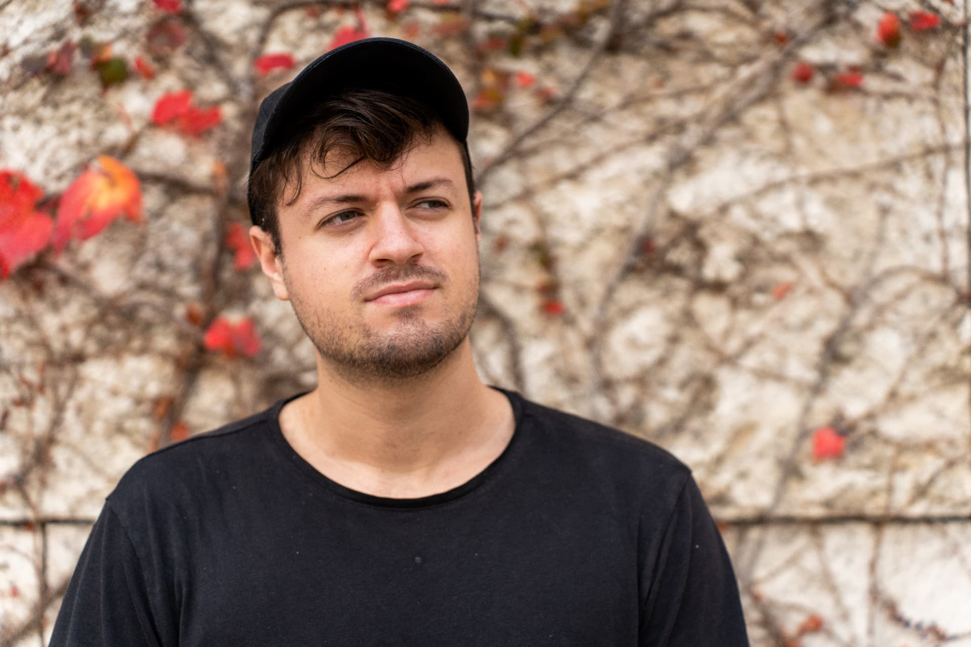 Portrait of a man in a black cap and tee against an ivy-covered stone wall with red autumn leaves