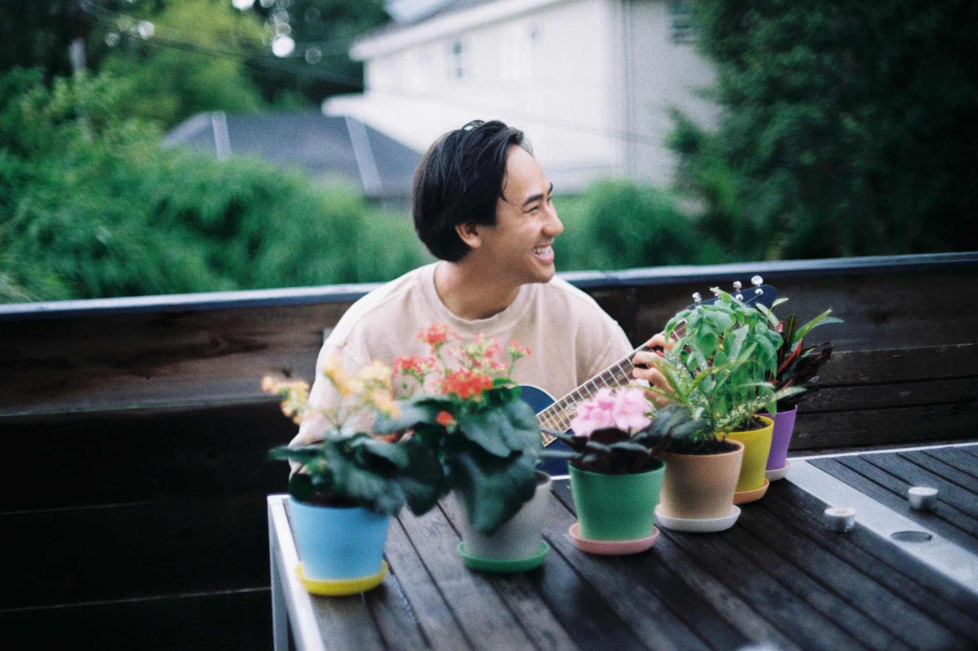 Film photo of a man laughing while playing ukulele on a porch behind a row of potted flowers