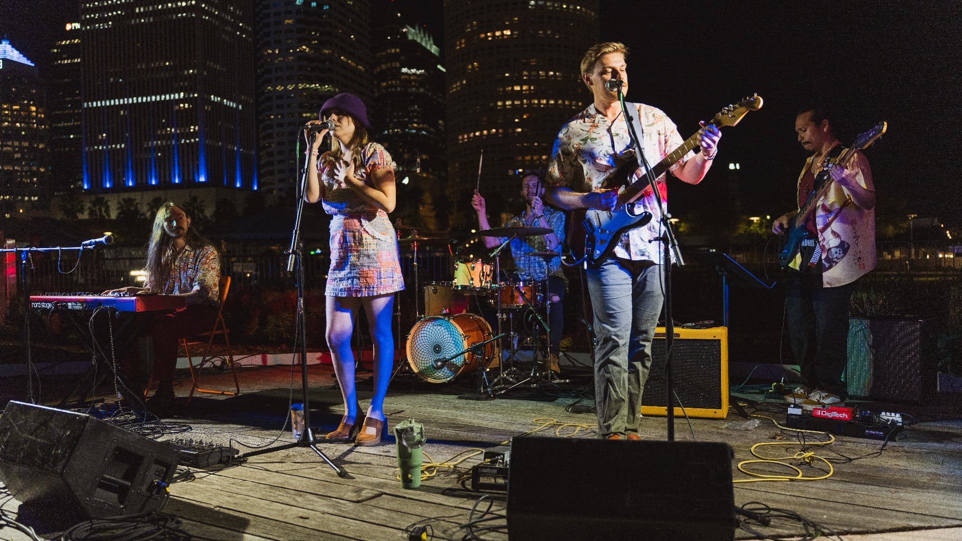 Mai performing as a full band on a waterfront stage with Tampa high-rises behind them
