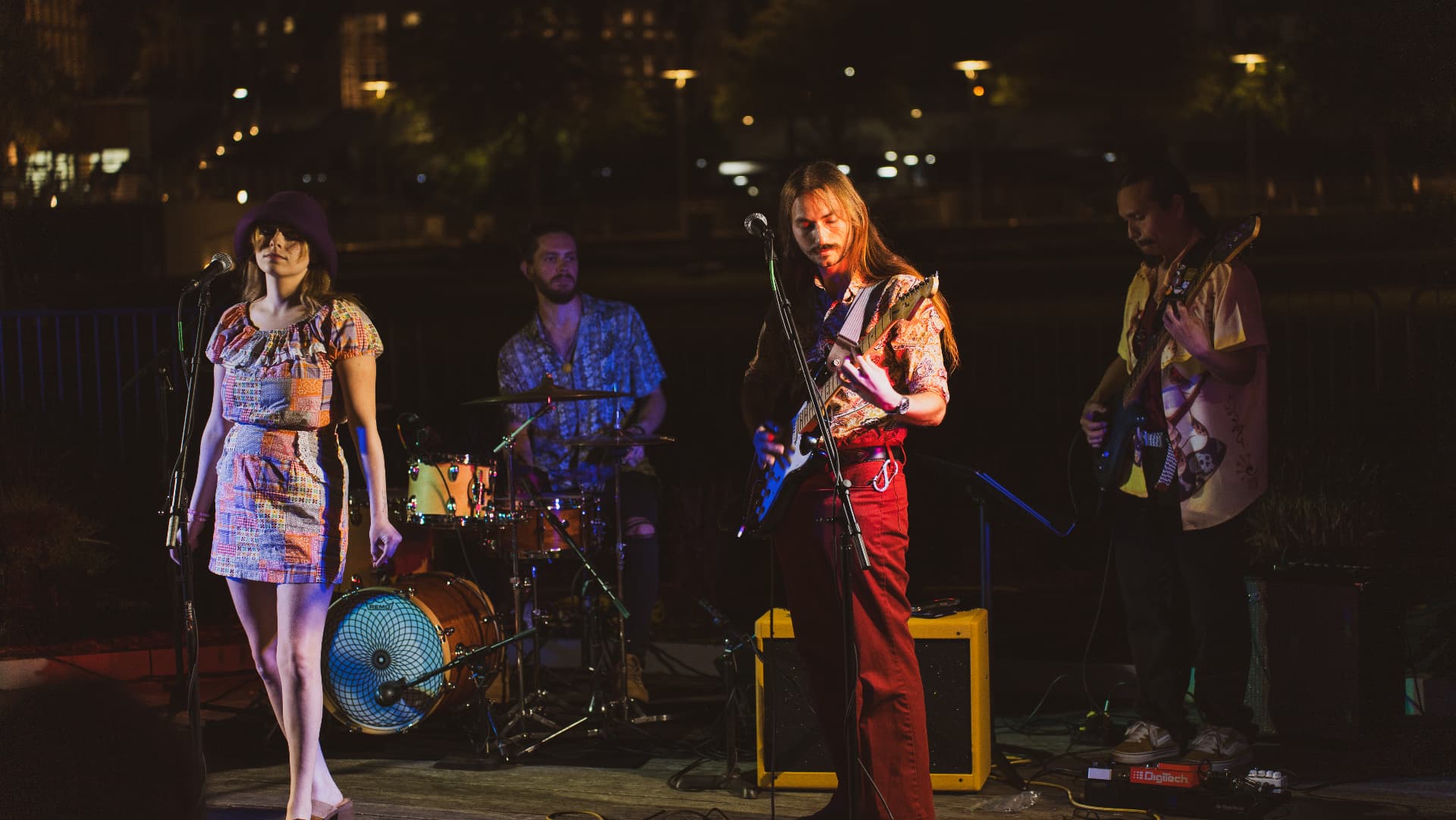 Mai's full band mid-set on an outdoor waterfront stage at night, city lights glowing behind them