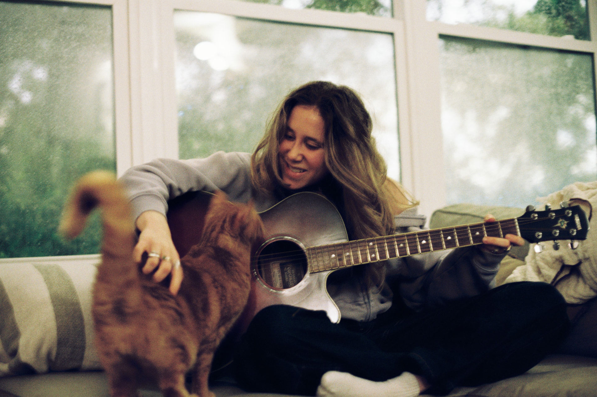 A woman laughing while playing acoustic guitar on a couch as her cat walks past