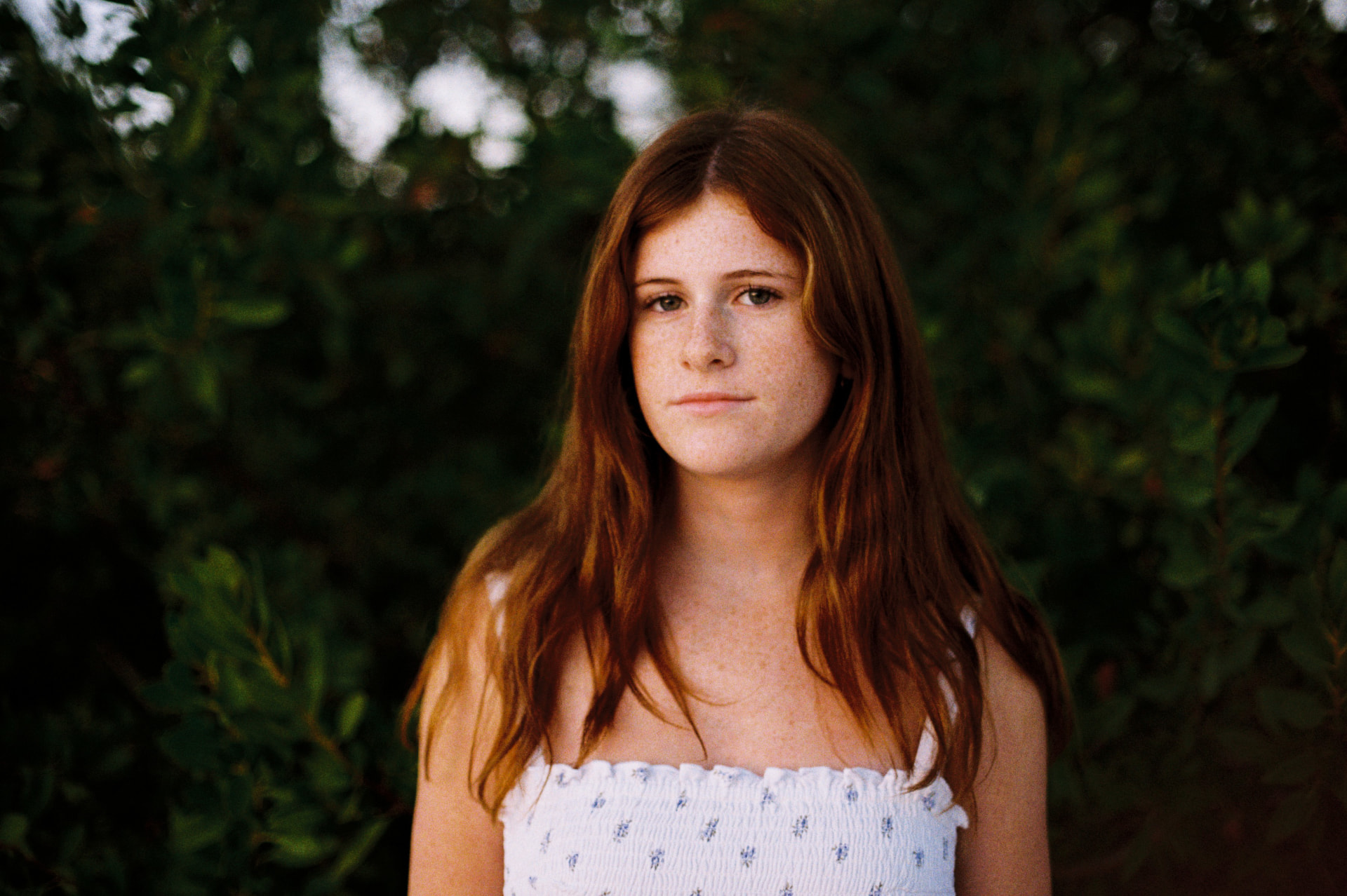 Golden-hour film portrait of a red-haired girl in front of dark green foliage