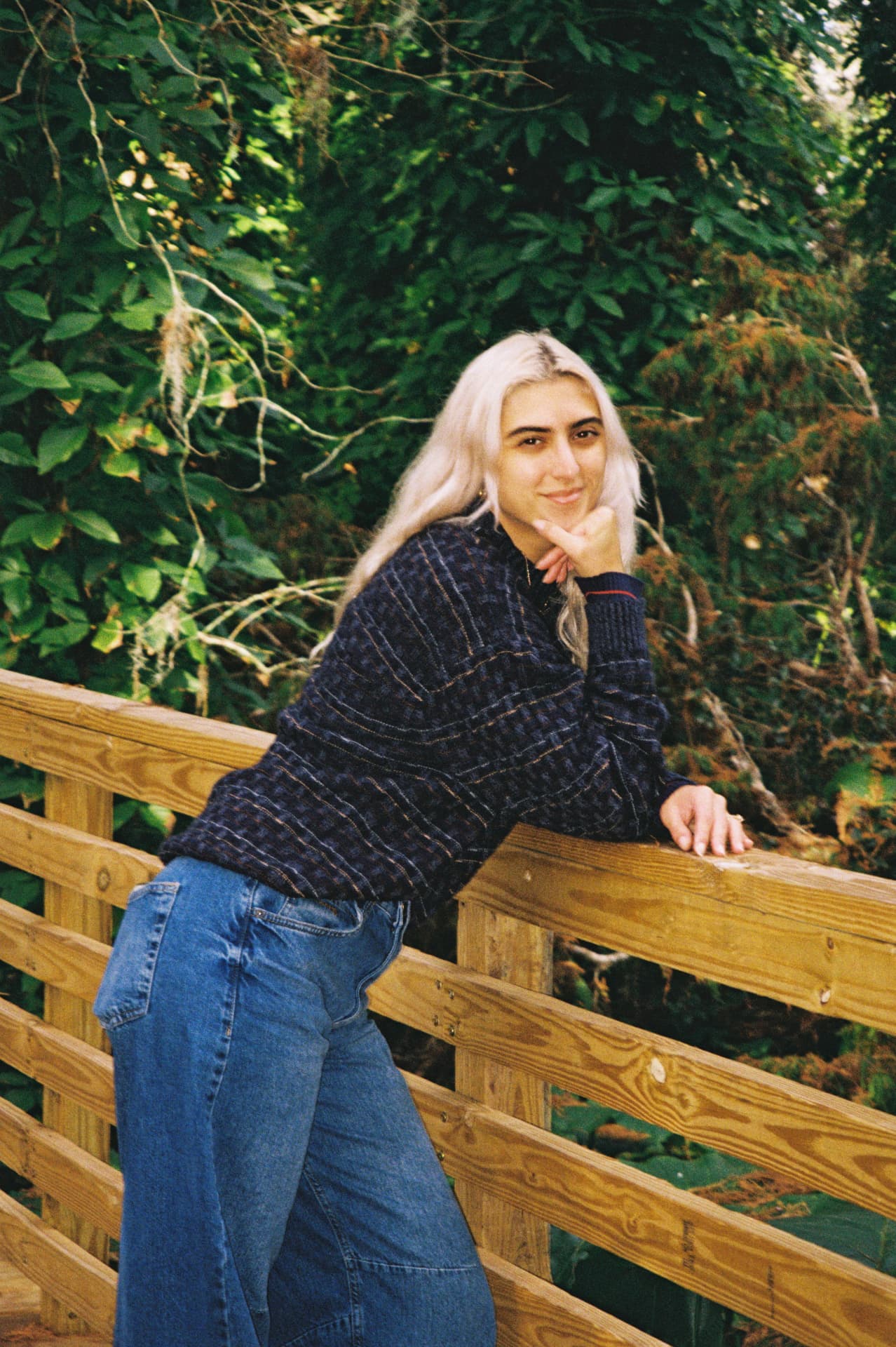 Film portrait of a woman with white hair leaning on a wooden bridge in lush greenery